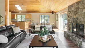 A modern living room with a leather sofa, a coffee table with a plant, a stone fireplace, and a kitchen in the background. - Tanner by Avantstay Modern Cascades Escape (Bend)
