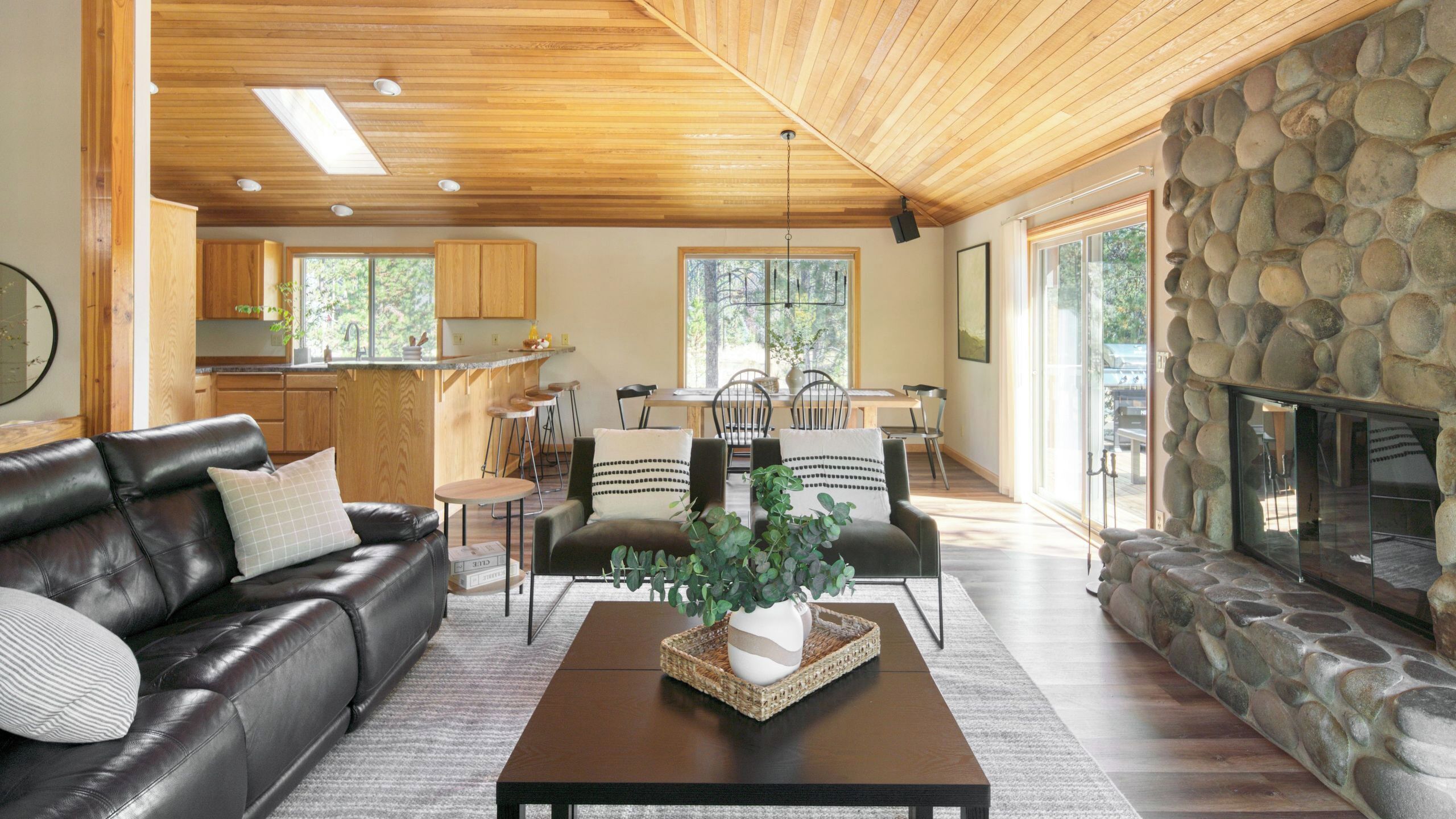 A modern living room with a leather sofa, a coffee table with a plant, a stone fireplace, and a kitchen in the background.
