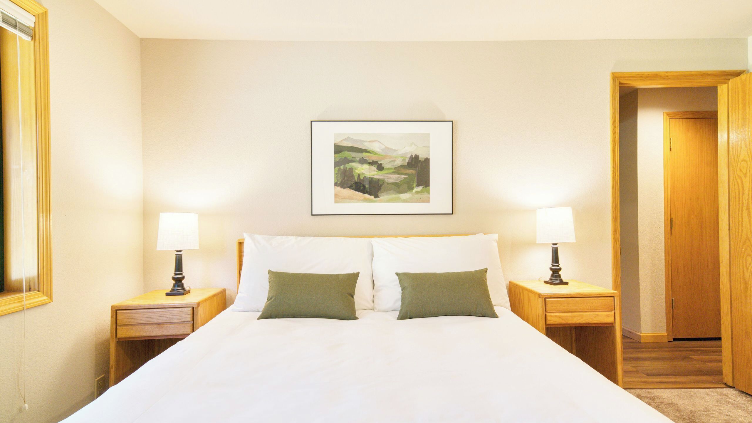 A hotel room with a large bed, two bedside tables with lamps, a framed landscape painting on the wall, and a wooden door.