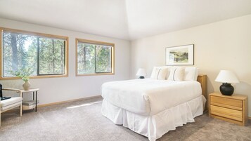 Ein Schlafzimmer mit einem großen Bett, zwei Nachttischen, einem Sessel, einem kleinen Tisch mit einer Pflanze und zwei Fenstern mit Blick auf einen Wald.
