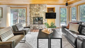 A living room with a stone fireplace, leather sofas, a flat-screen TV, and large windows. - Tanner by Avantstay Modern Cascades Escape (Bend)