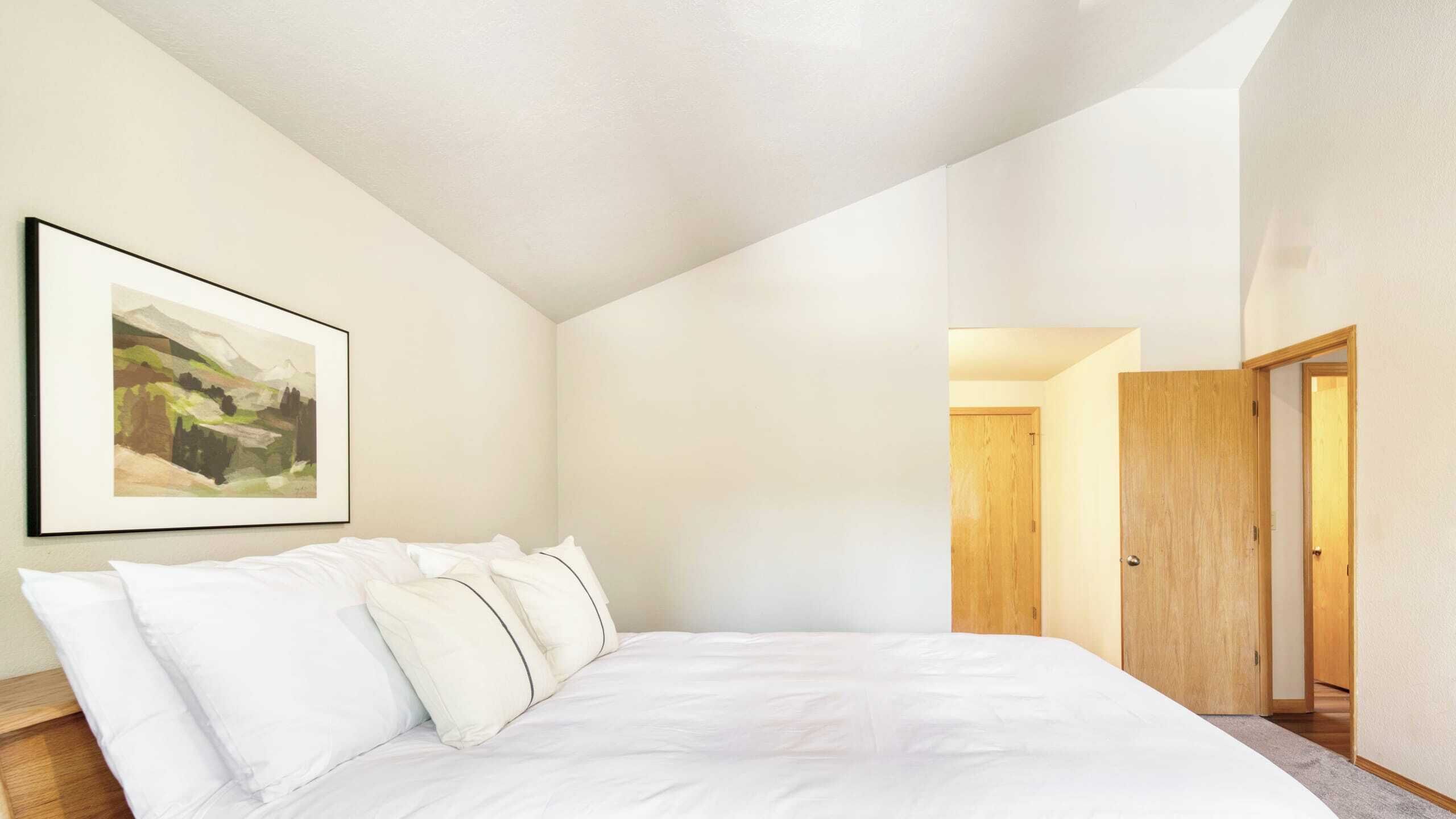 A bedroom with a large bed, a framed landscape painting, and a wooden door.