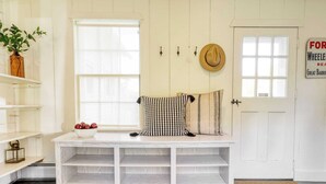 Un intérieur blanc avec un banc, un saladier de pommes, un chapeau accroché au mur et un paillasson sur lequel est écrit « AVANTSTAY ».