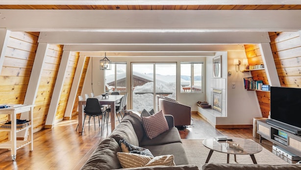 A cozy loft-style living room with a sofa, coffee table, and TV, featuring wooden walls and large windows overlooking a snowy landscape.
