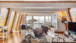 Un salon chaleureux de style loft, avec un canapé, une table basse et un téléviseur, agrémenté de murs en bois et de grandes fenêtres donnant sur un paysage enneigé.