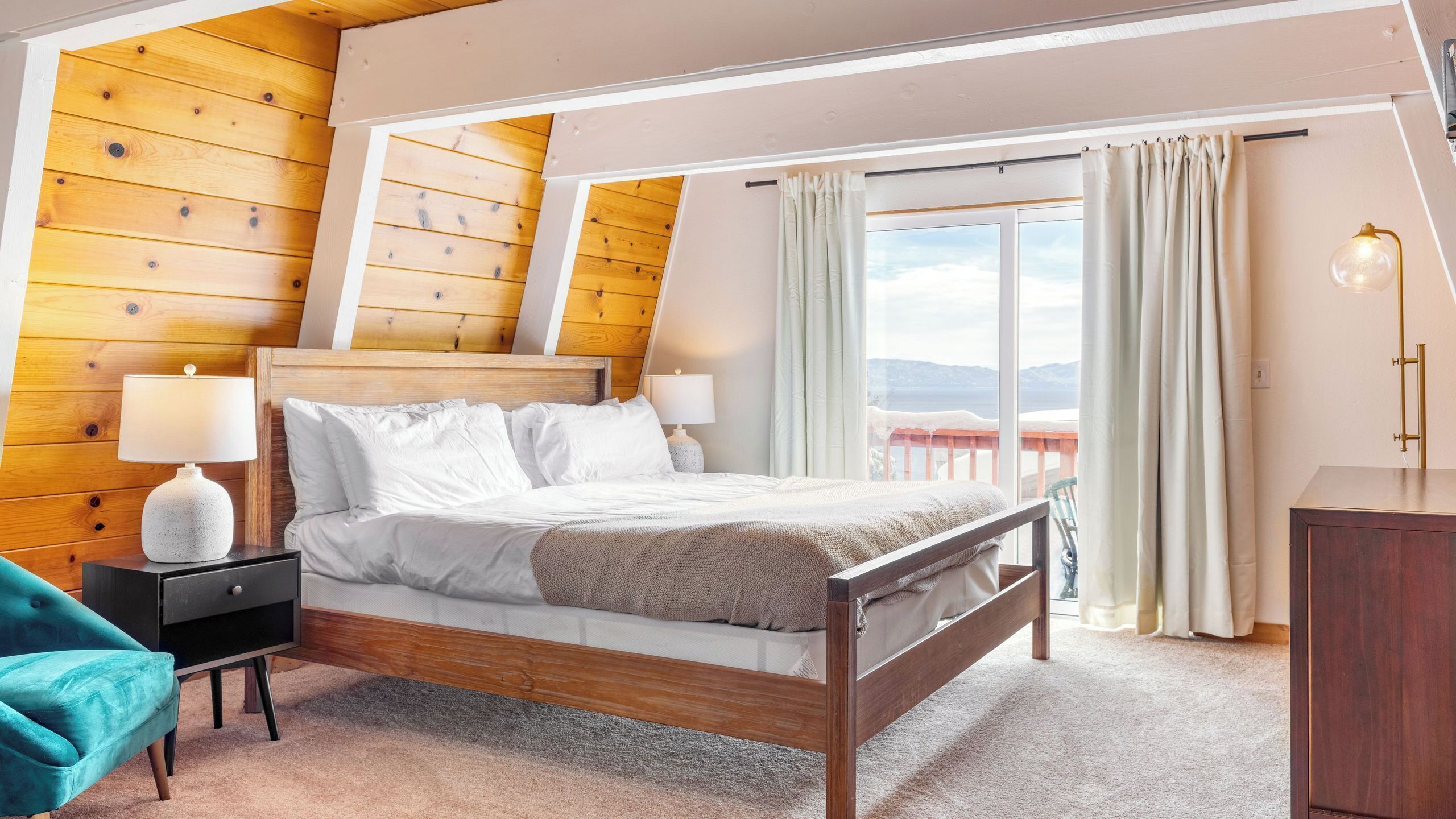 Un dormitorio con techo de madera, una cama con ropa de cama blanca, dos lámparas de noche, un sillón azul aguamarina y vistas al exterior.