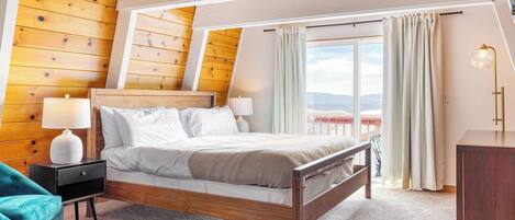 Un dormitorio con techo de madera, una cama con ropa de cama blanca, dos lámparas de noche, un sillón azul aguamarina y vistas al exterior.