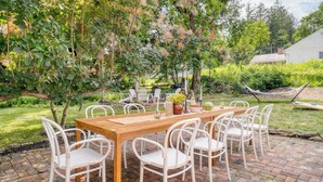 An outdoor dining area with a wooden table and white chairs, surrounded by lush greenery and trees. - Cordelia by Avantstay Amazing Backyard, 10mins From Stockbridge! (Lee)