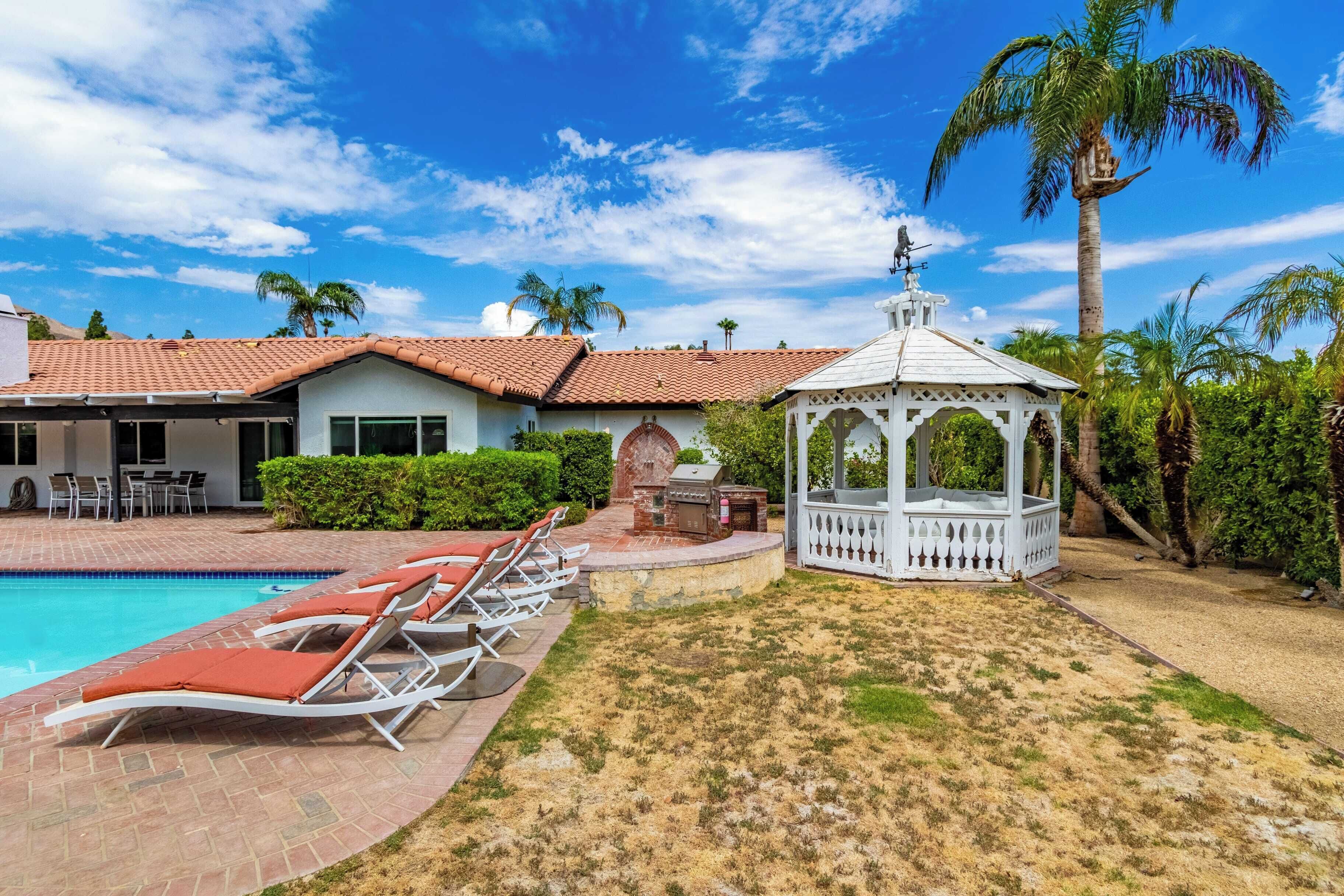 A pool area with lounge chairs, a gazebo, and a brick building with a tiled roof.