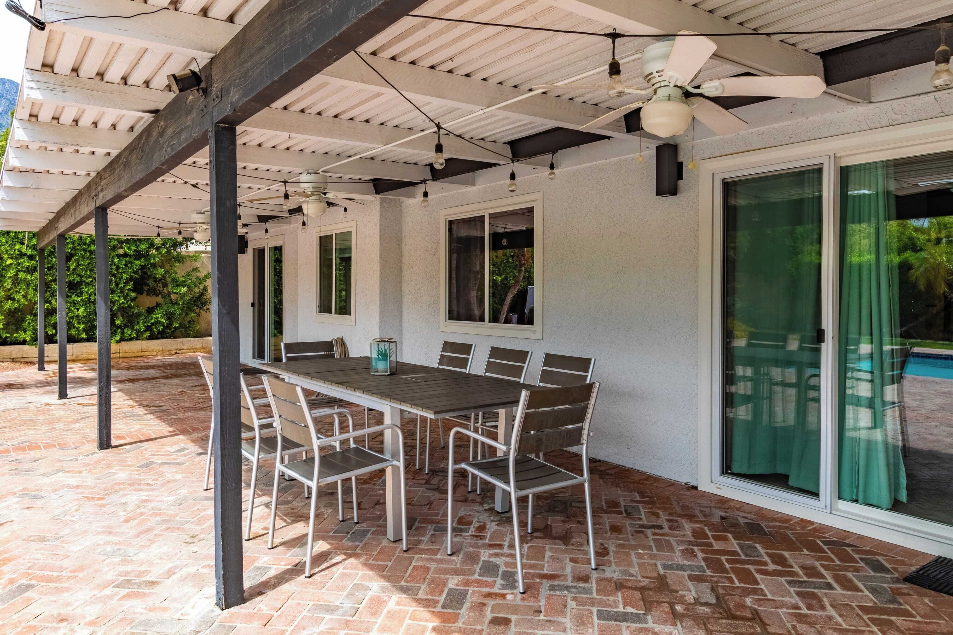 An outdoor patio with a brick floor, a wooden table with metal chairs, a ceiling fan, and a glass door leading to a pool.