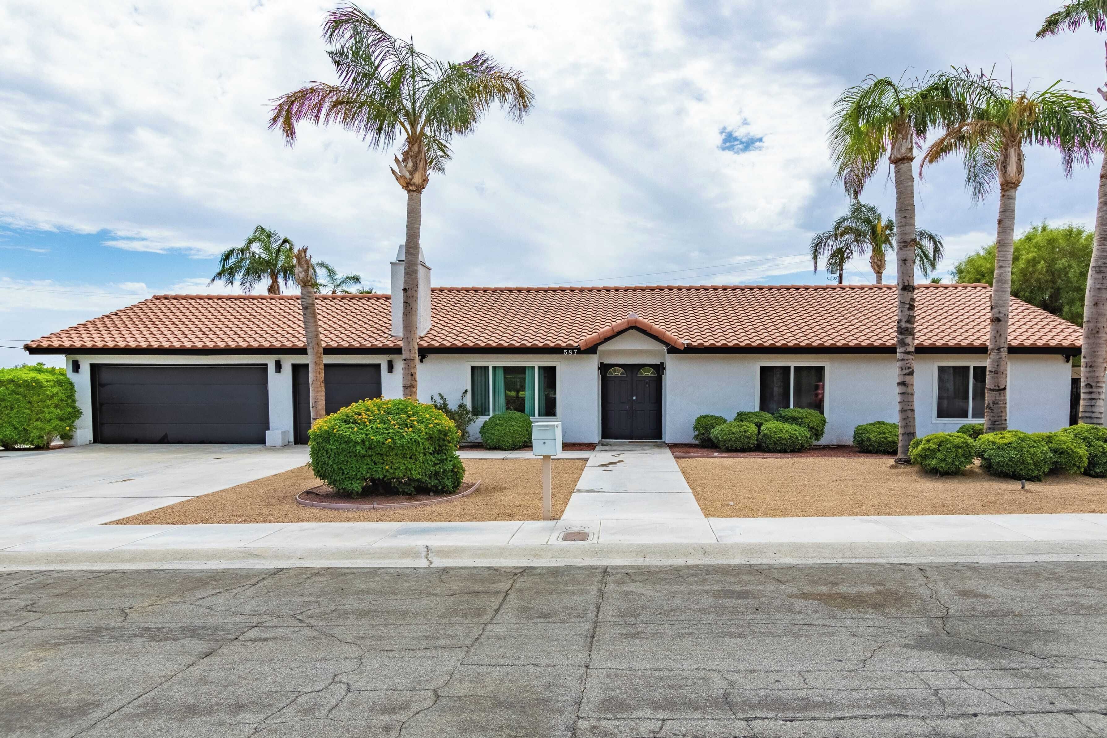 A two-story house with a red-tiled roof, a garage, and a front yard with palm trees and shrubs.