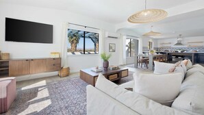 A spacious living area with a large sofa, a coffee table, and a TV mounted on the wall. There is a large window providing a view of palm trees and mountains.
