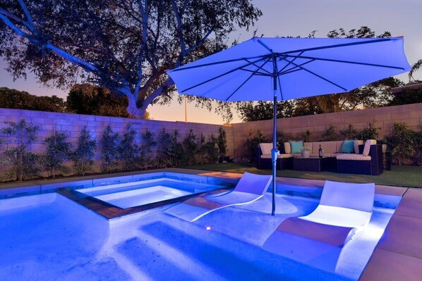 A swimming pool with a blue-lit deck, lounge chairs, and a patio area with outdoor furniture.