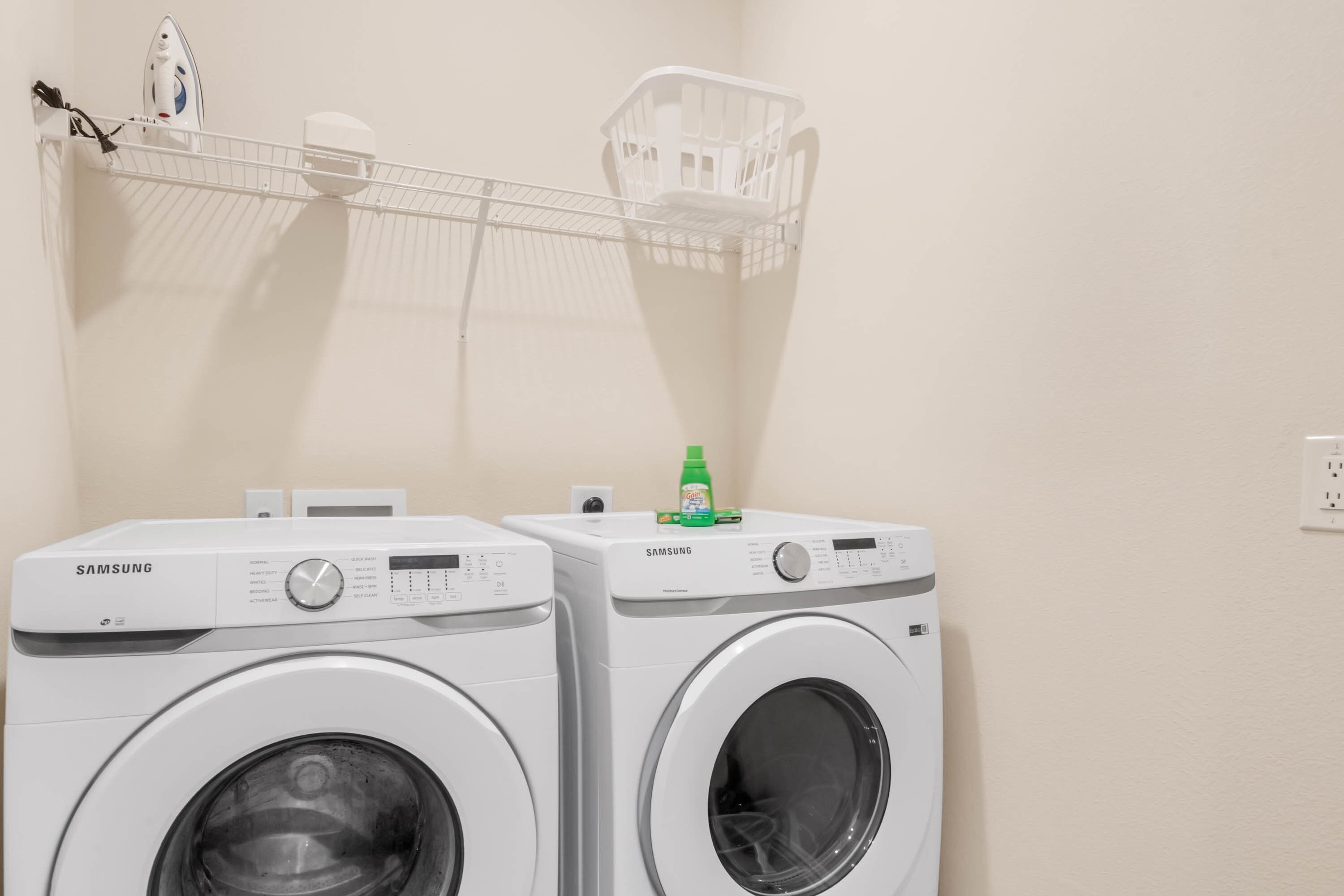 Deux machines à laver Samsung à chargement frontal sont installées dans un espace dédié à la buanderie, avec un mur blanc, un panier à linge et une bouteille de lessive Gain posée sur le dessus.