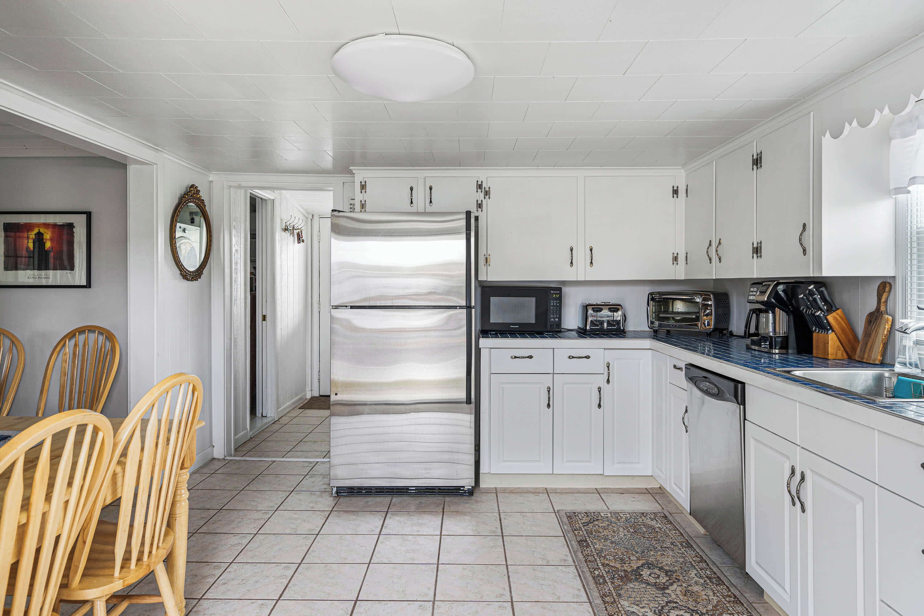 A kitchen with white cabinets, a stainless steel refrigerator, a microwave, and a toaster. There is a dining table with chairs and a rug on the floor.