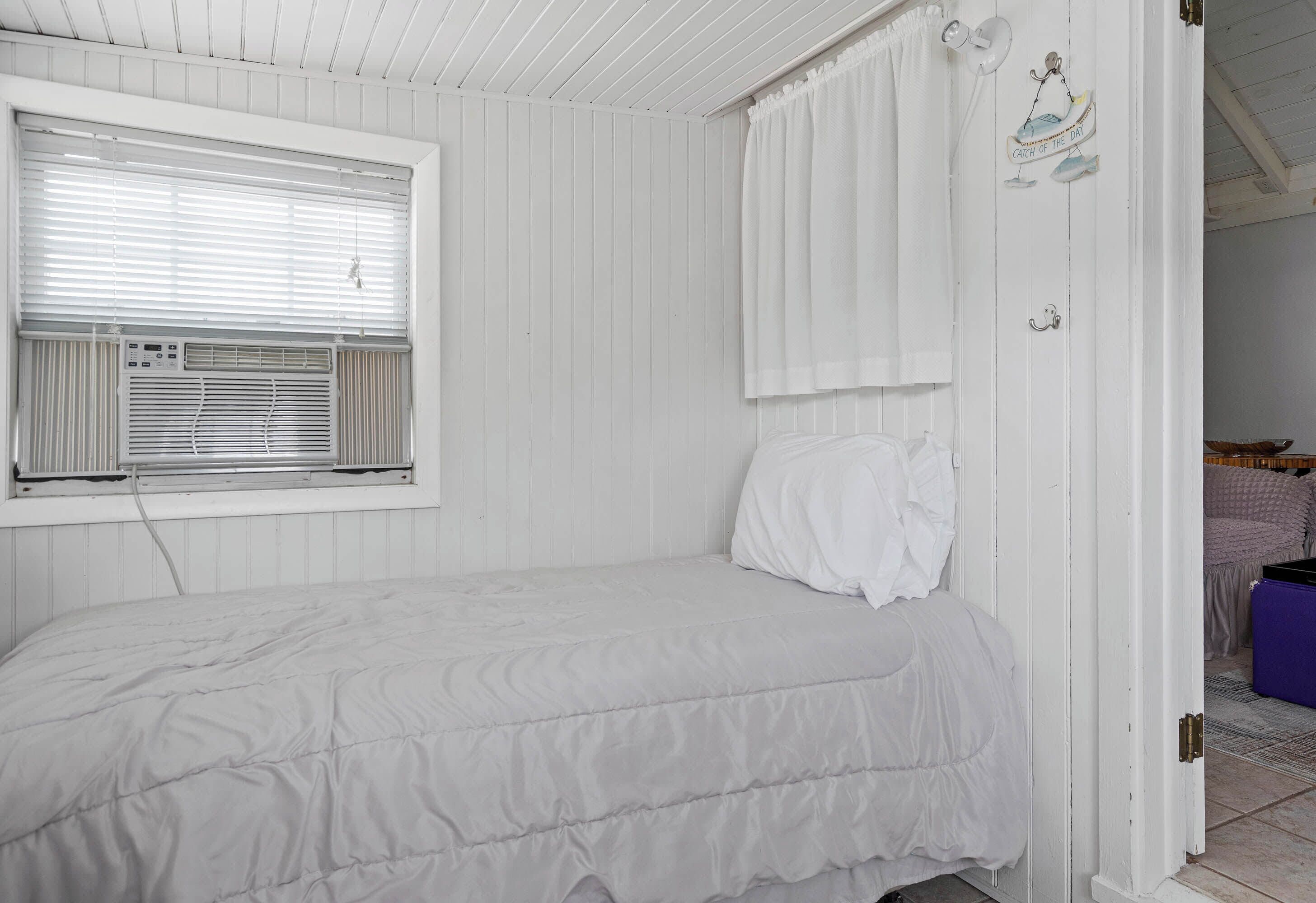Quarto branco com uma cama, janela com persianas, porta com cortina e ar condicionado fixado na parede.