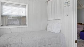 Quarto branco com uma cama, janela com persianas, porta com cortina e ar condicionado fixado na parede.