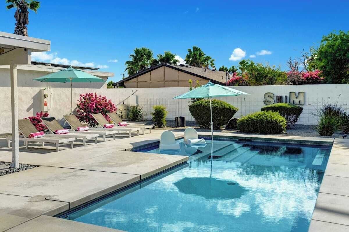 Una piscina con sillones y sombrillas, rodeada por un jardín ajardinado y un edificio con el letrero "SIM".