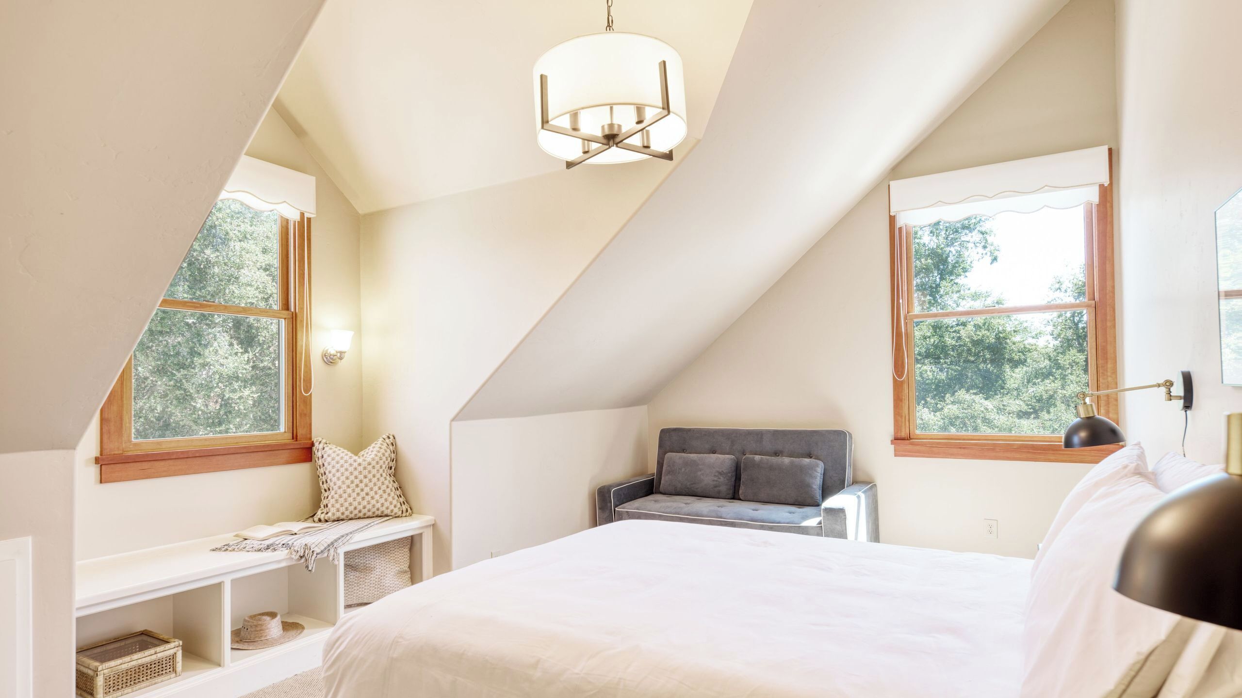Un dormitorio con techo inclinado, una cama, un banco y dos ventanas con vista a árboles.