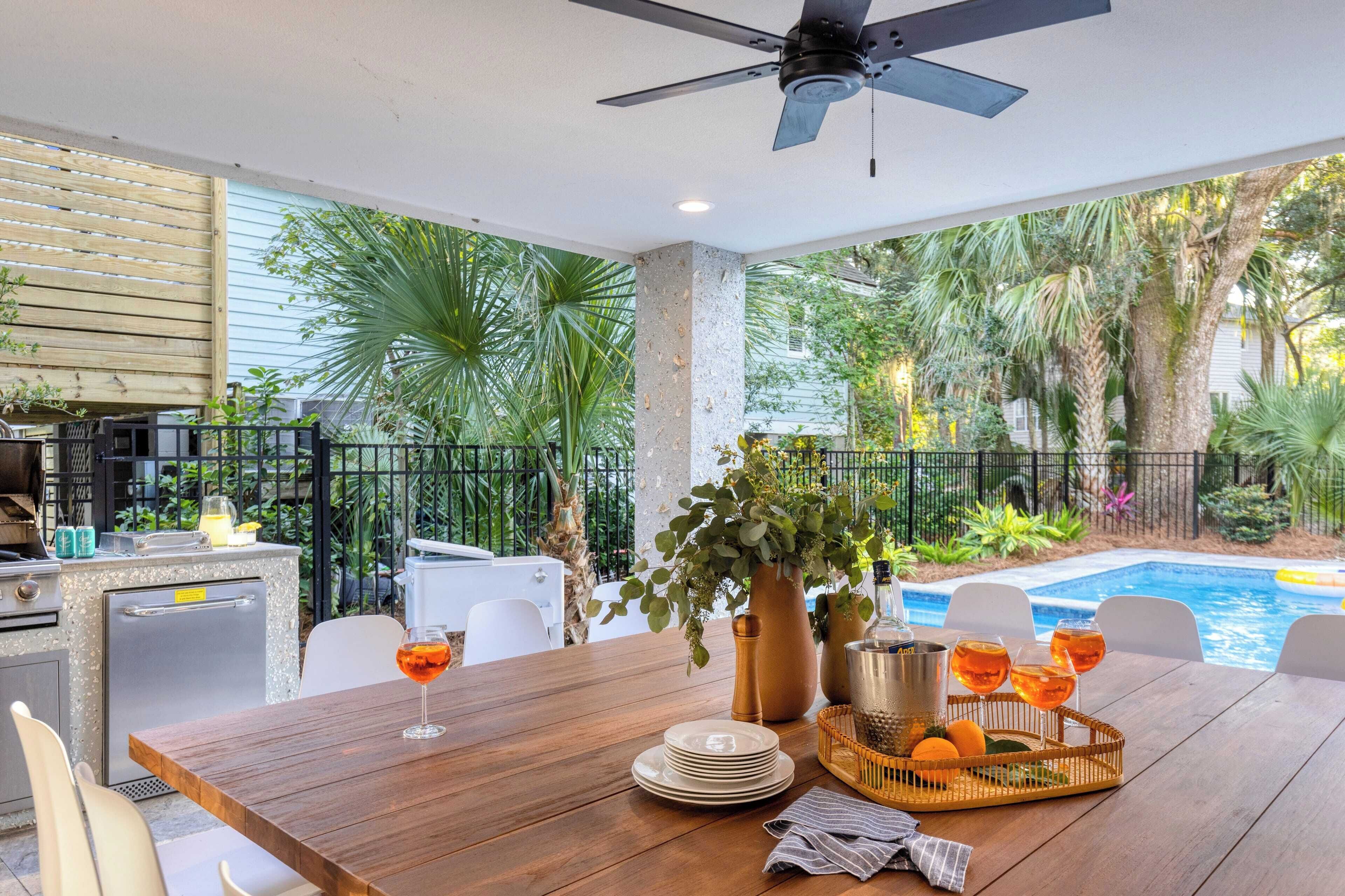 An outdoor dining area with a wooden table, chairs, a ceiling fan, a pool, and lush greenery.
