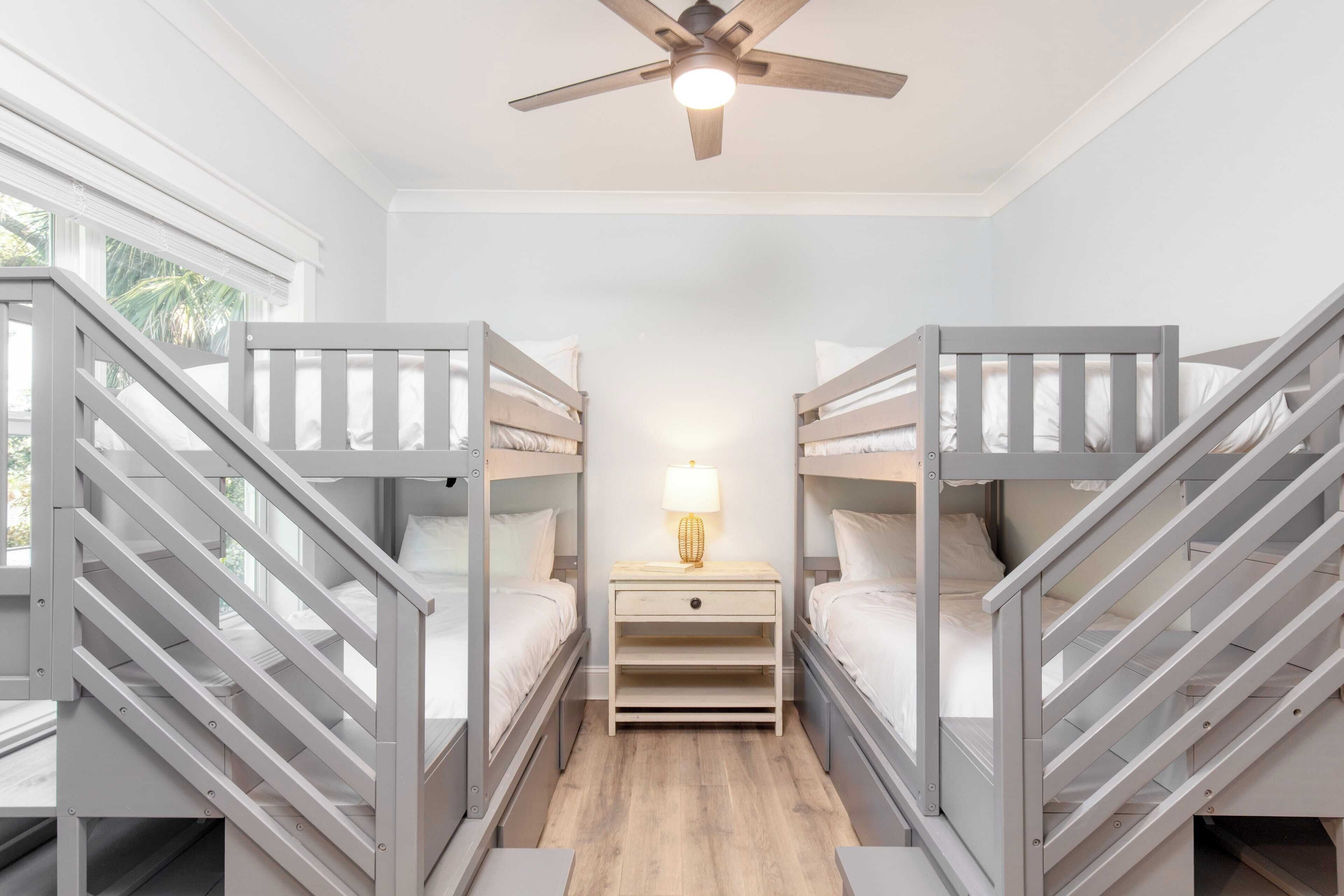 A room with bunk beds, a ceiling fan, a nightstand with a lamp, and a window with a view of palm trees.