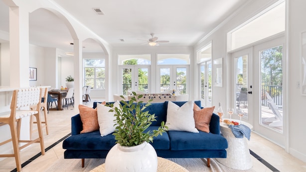 A spacious living room with a blue sofa, white and orange cushions, and a large white vase with green plants. - Miramar Manor by Avantstay Walk to the Beach! (Miramar Beach)