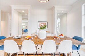 A dining area with a wooden table, white chairs, a vase with flowers, and a bowl of fruit. - Miramar Manor by Avantstay Walk to the Beach! (Miramar Beach)
