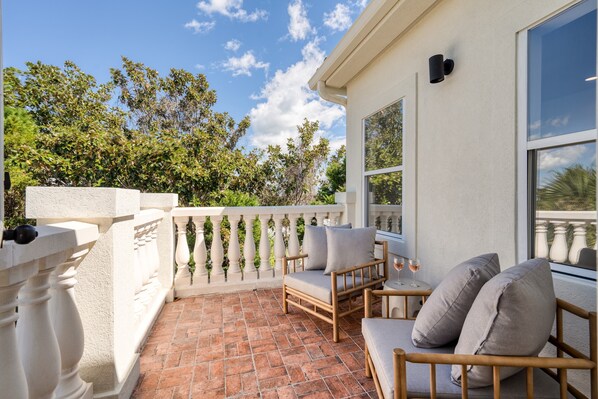 A balcony with a white balustrade, a wooden sofa with cushions, and a table with two wine glasses.