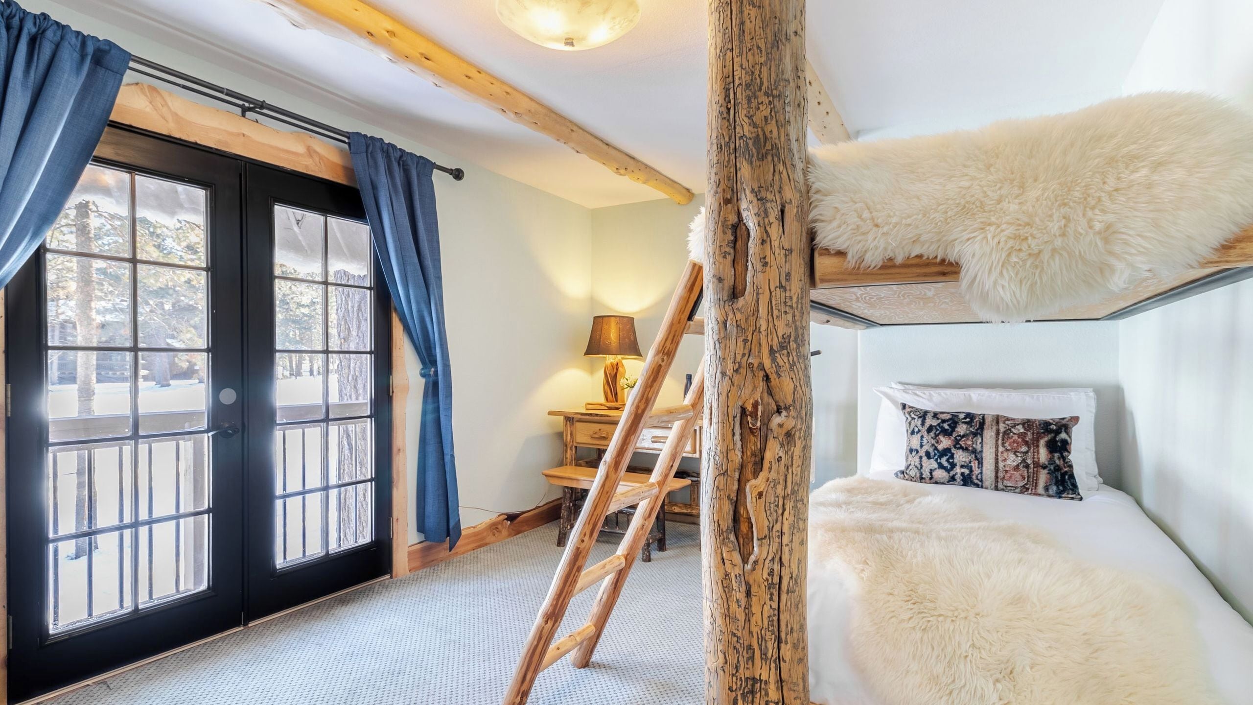Une chambre avec un lit superposé, un bureau avec une lampe, une échelle et une grande fenêtre avec des rideaux.