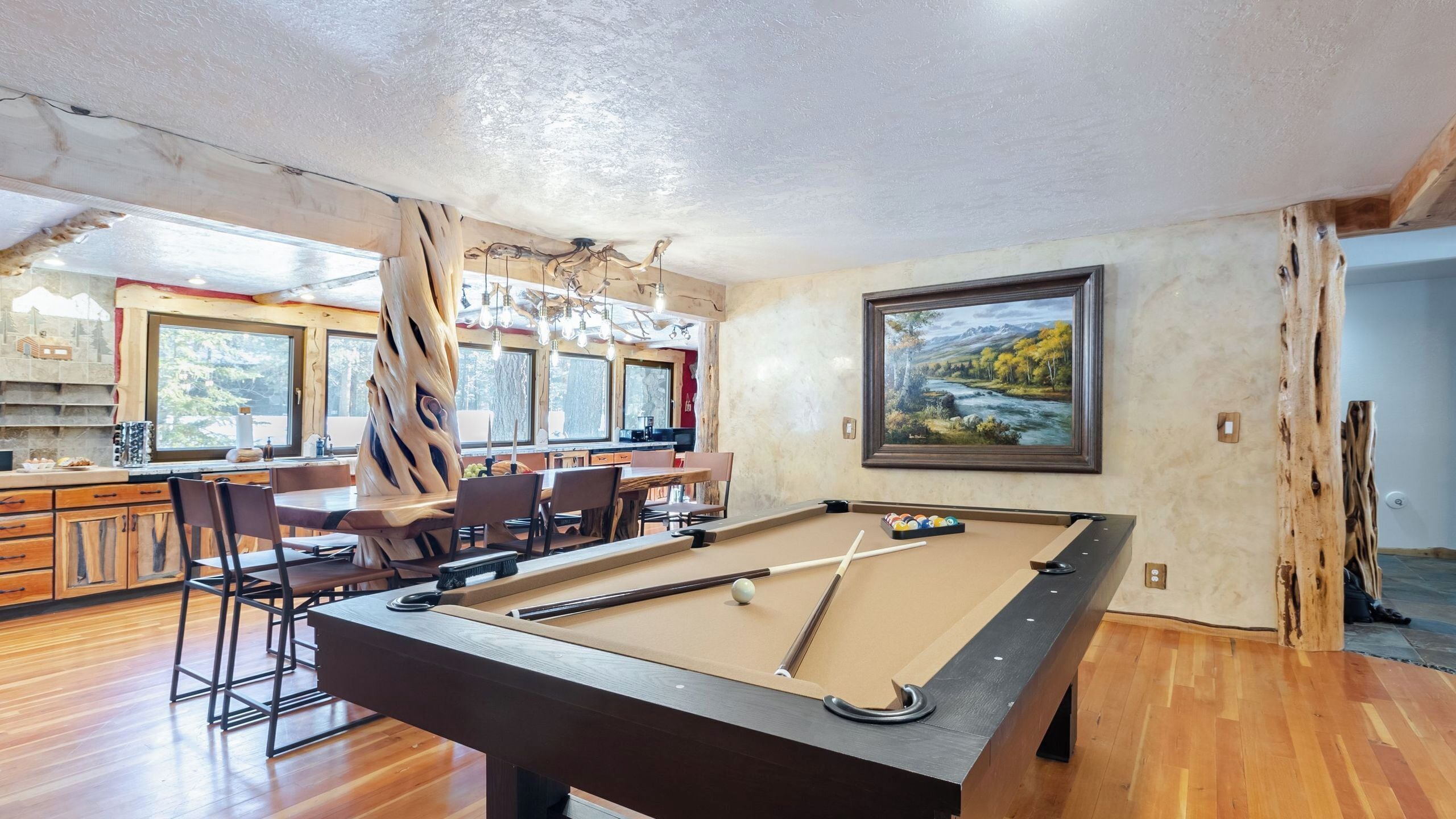 A pool table in a room with wooden flooring, a dining area with chairs and a table, and a painting on the wall.