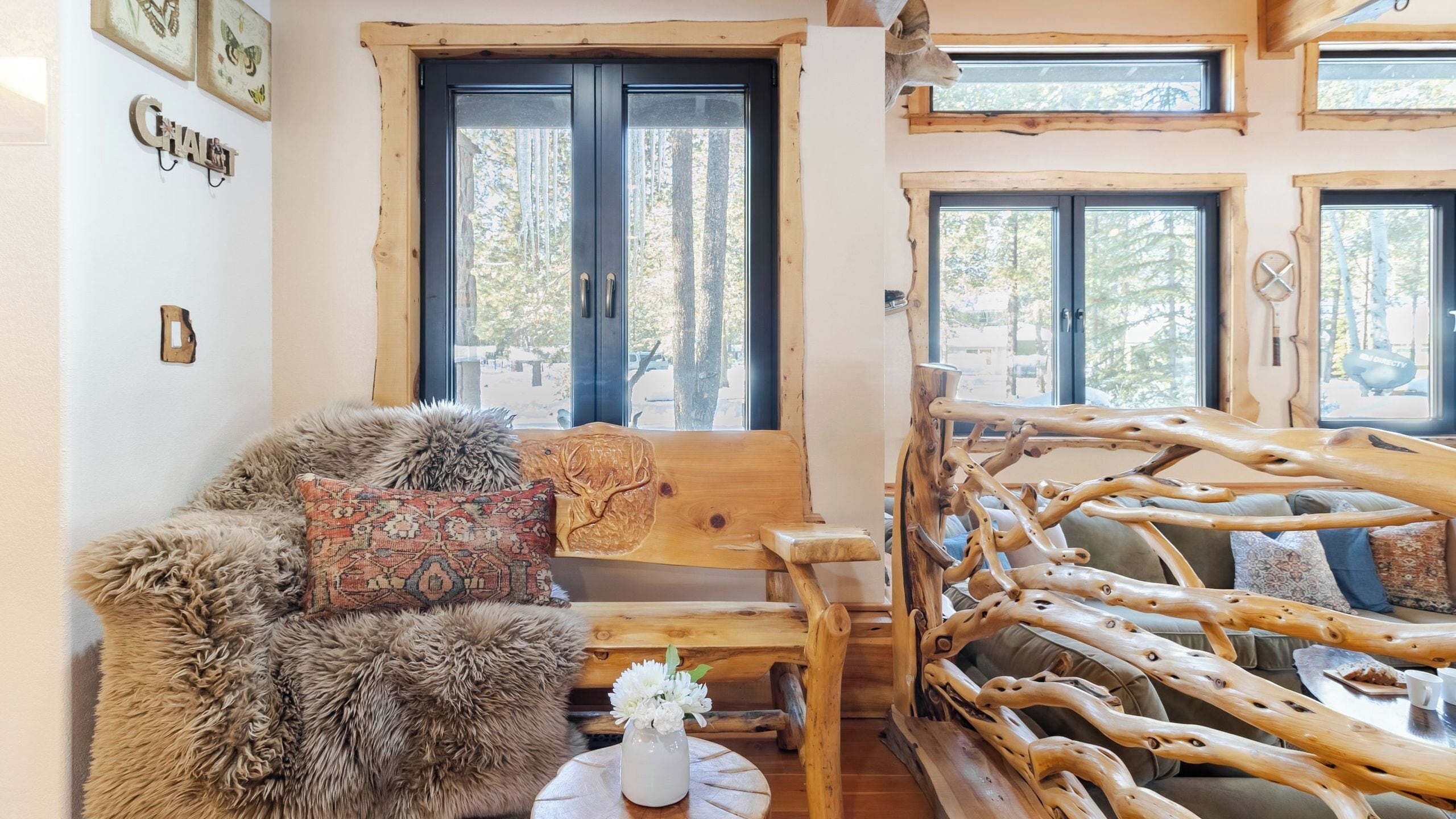 A rustic cabin interior with a wooden bench, a fur-covered chair, and a wooden table with a vase of flowers.