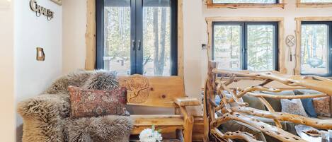 A rustic cabin interior with a wooden bench, a fur-covered chair, and a wooden table with a vase of flowers.