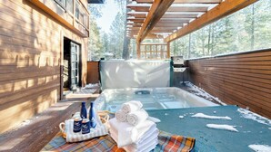 A hot tub area with a wooden deck, a covered seating area, and a view of the surrounding forest. - Base Camp by Avantstay Magical Cabin w/ Sauna, Hot Tub & Close to Snow & Golf (Bend)