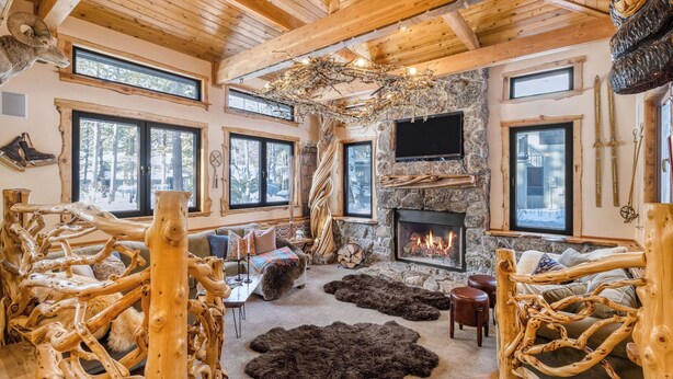A log cabin interior with a stone fireplace, wooden beams, and a large window overlooking a forest. - Base Camp by Avantstay Magical Cabin w/ Sauna, Hot Tub & Close to Snow & Golf (Bend)