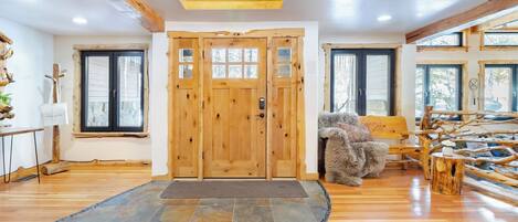 A wooden door with glass panels, a stone-tiled entrance mat, a bench with a fur throw, and a rustic wooden wall with a window.
