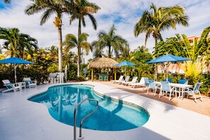 A swimming pool with lounge chairs and umbrellas, surrounded by palm trees and a thatched-roof structure.