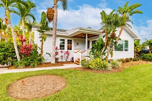 A white house with a porch, surrounded by tropical plants and a well-maintained lawn.