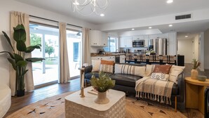 A modern living room with a sofa, coffee table, and potted plants. There is a kitchen in the background with stainless steel appliances.
