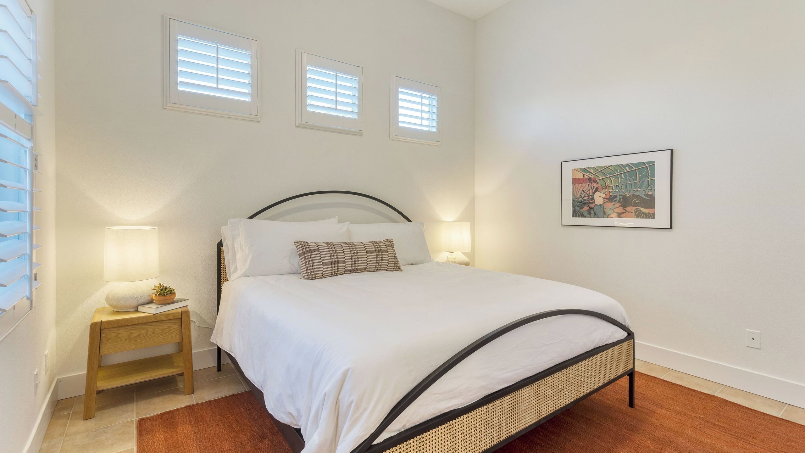 Ein ordentlich eingerichtetes Schlafzimmer mit einem Bett, Nachttischlampen, einem kleinen Tisch und einem gerahmten Bild an der Wand.
