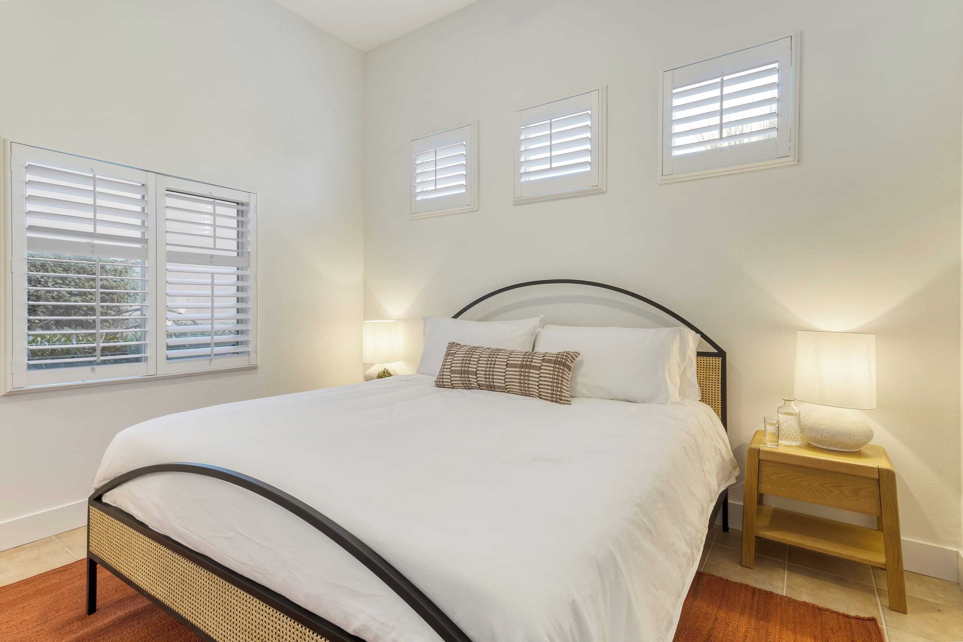A neatly arranged bedroom with a bed, bedside lamps, and three windows with blinds.