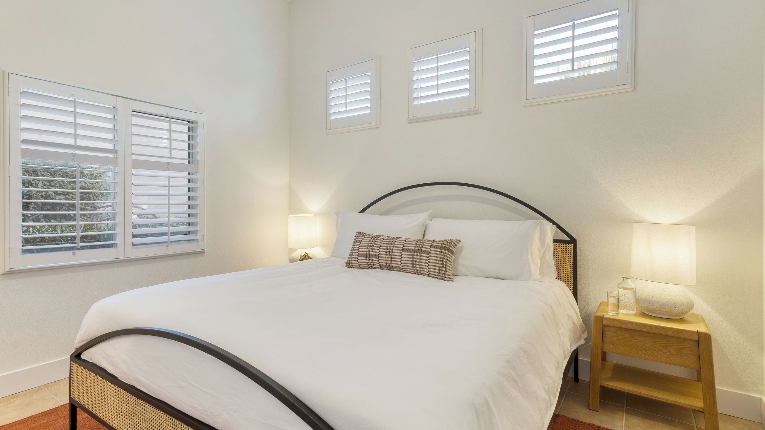 A bedroom with a bed, bedside lamps, and three windows with blinds.