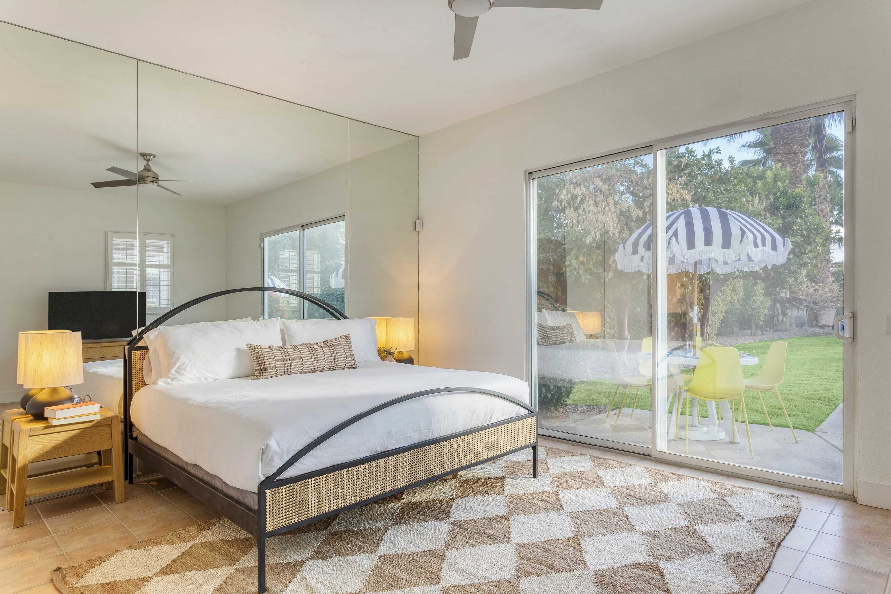 A bedroom with a bed, bedside lamps, a ceiling fan, a large mirror, and a sliding glass door leading to an outdoor area with chairs and an umbrella.