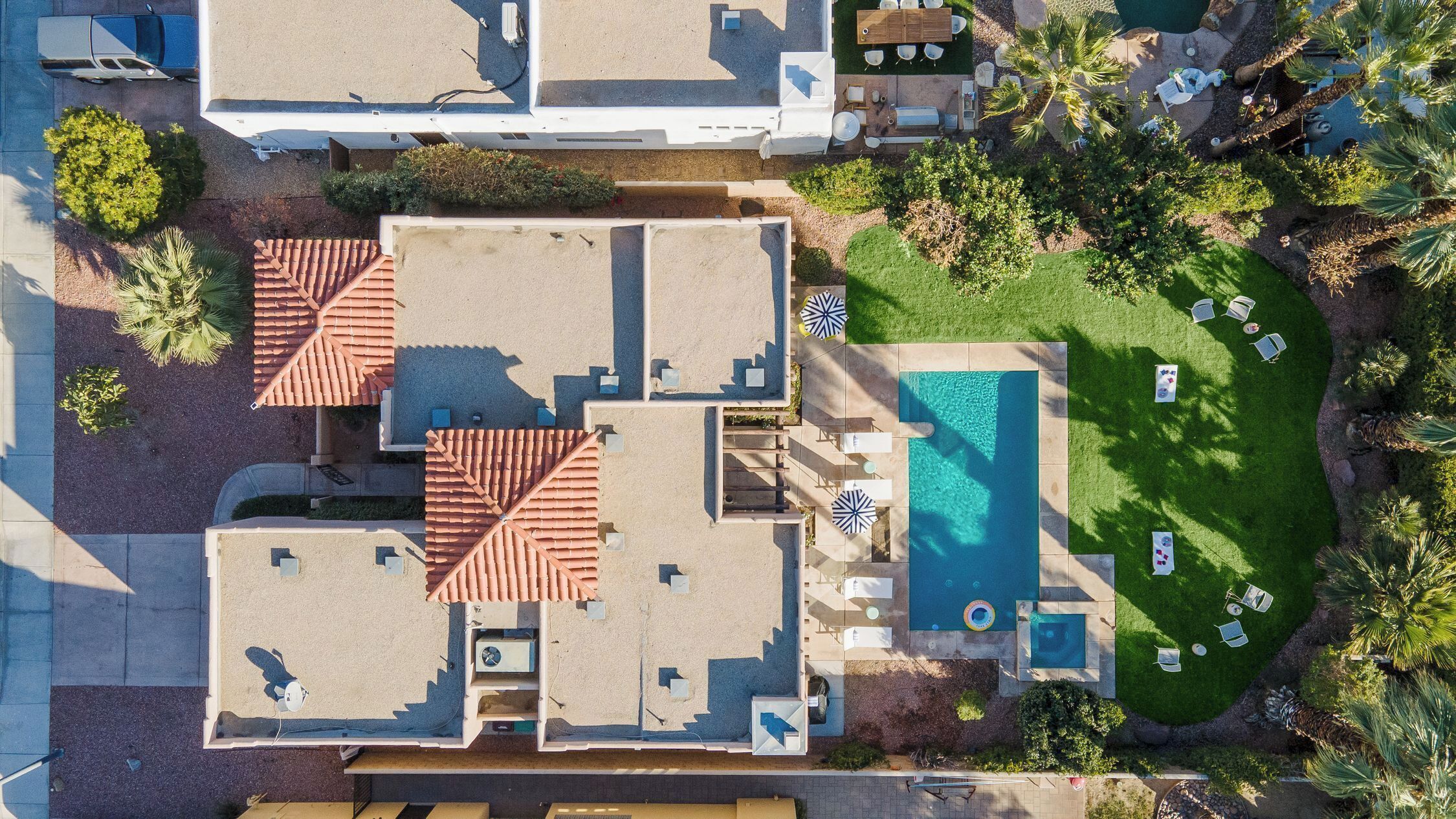 A rooftop pool with lounge chairs and umbrellas, surrounded by greenery and a parking area.