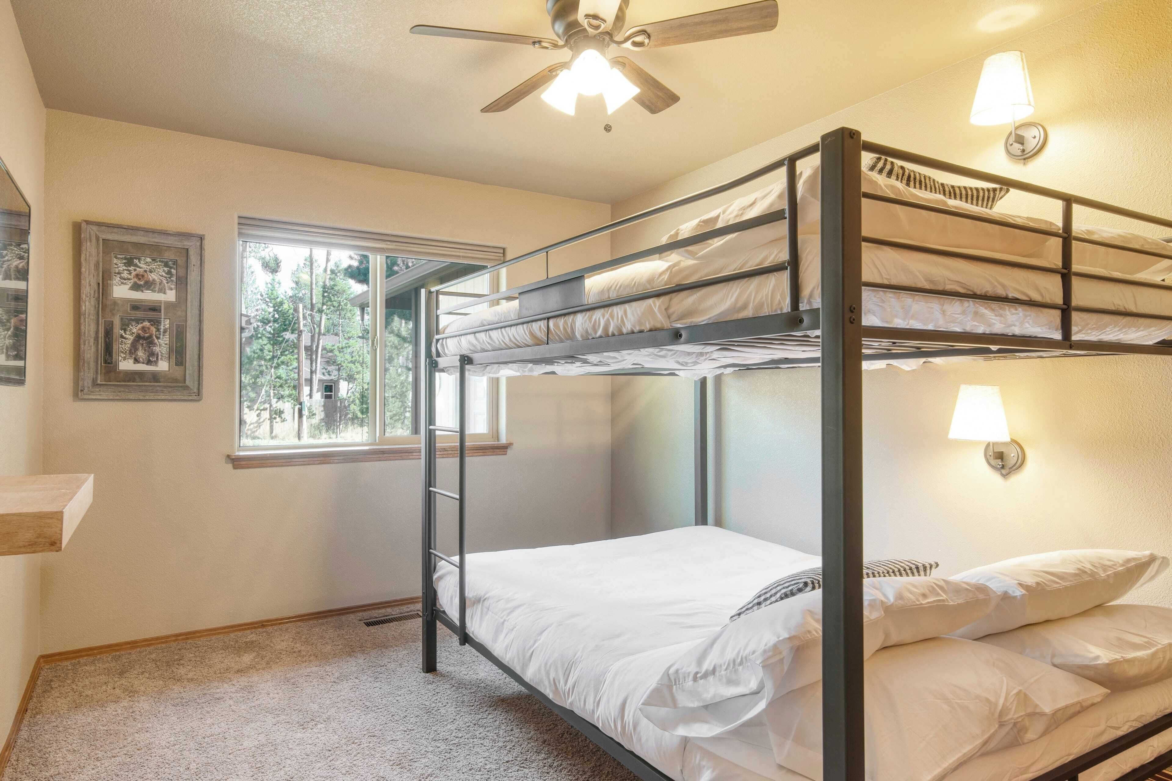 A bunk bed room with a ceiling fan, two wall-mounted lamps, a window with a view of trees, and a framed picture on the wall.