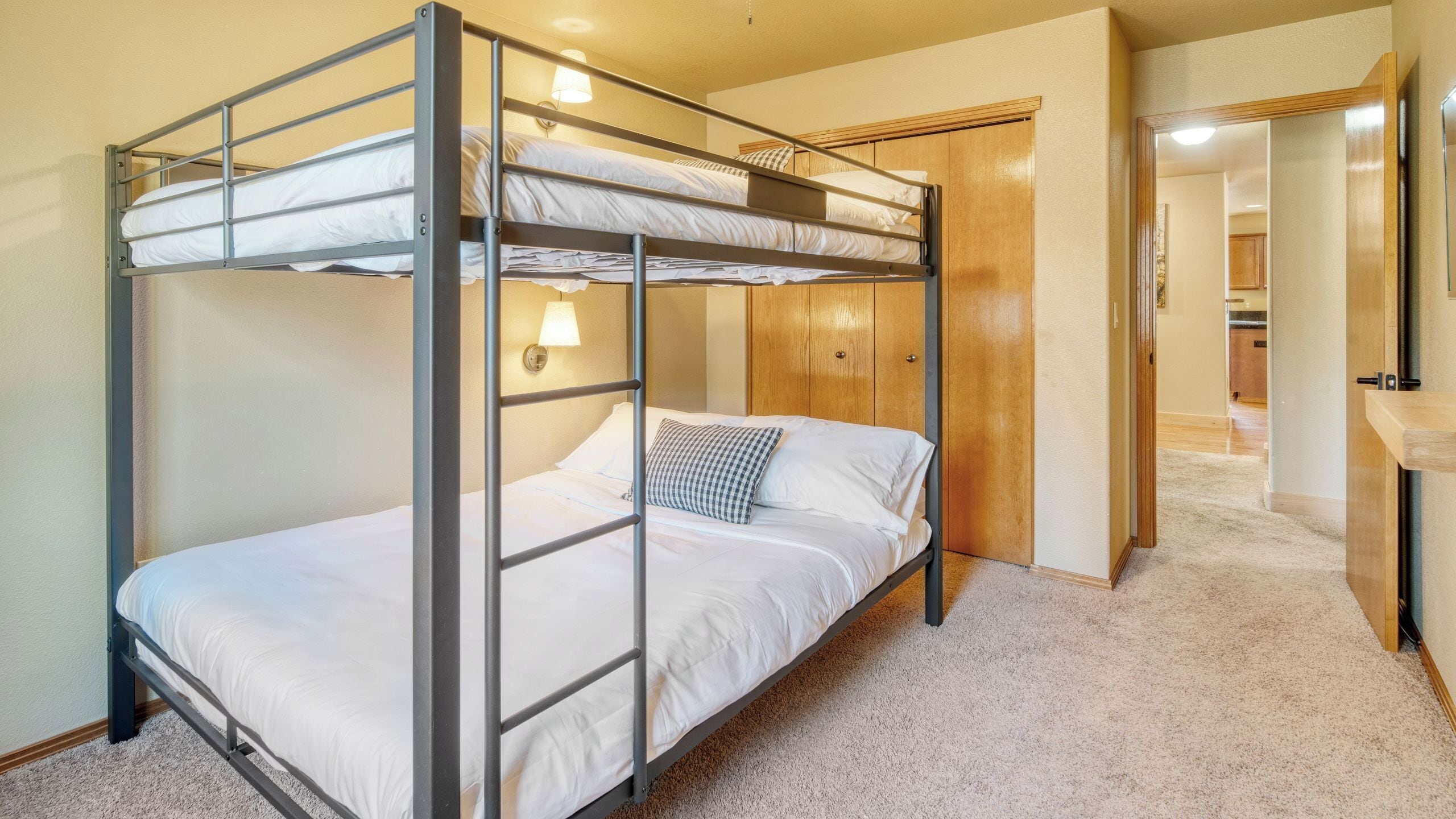 A bunk bed in a room with a carpeted floor, a wooden wardrobe, and a lamp.