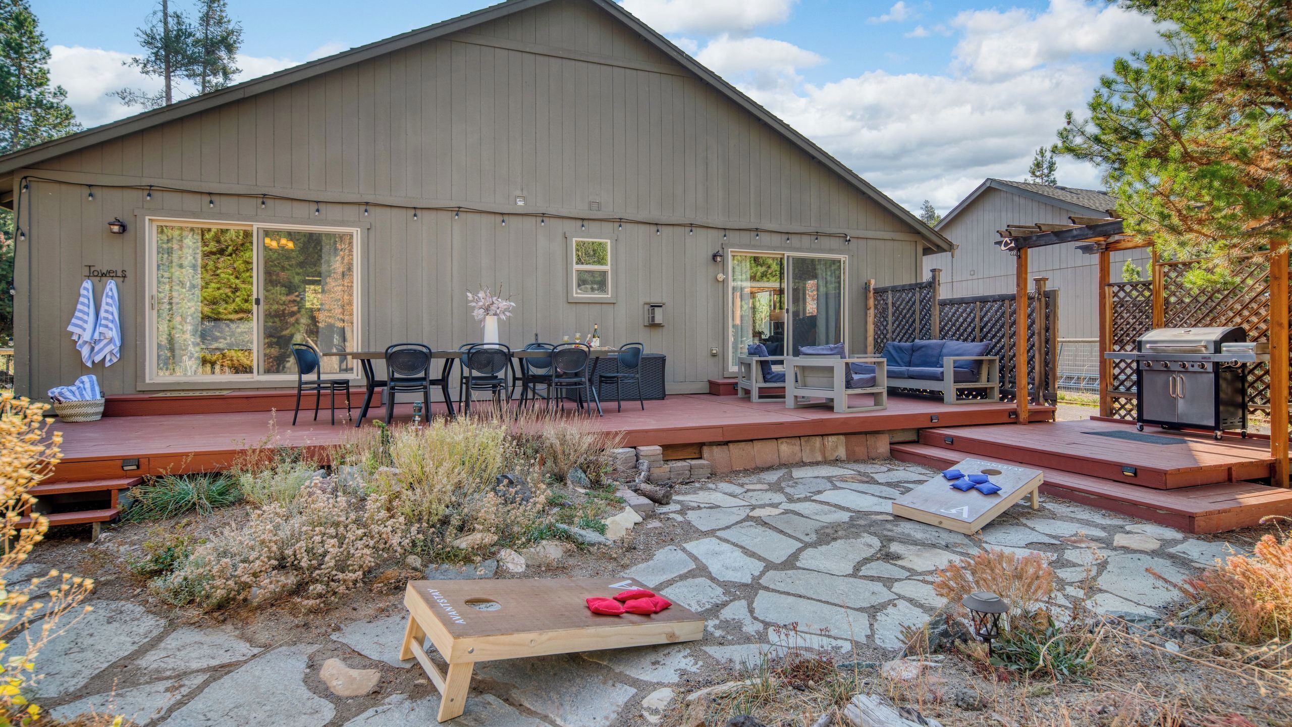 Un patio couvert avec des meubles d’extérieur, un barbecue et un chemin de pierres menant à une terrasse en bois.