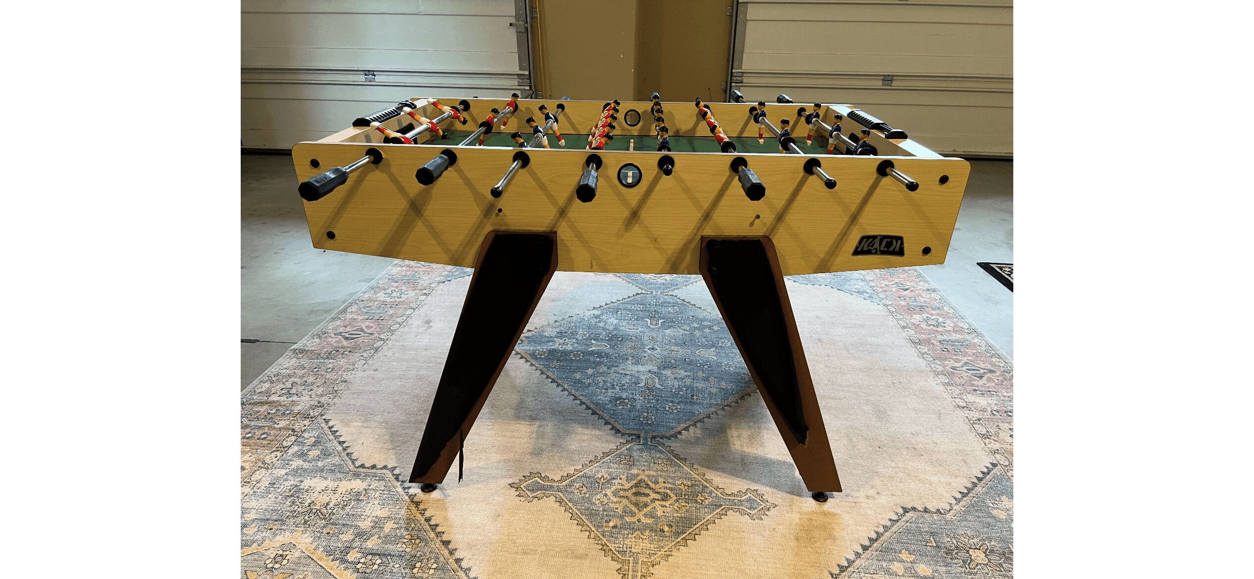 Una mesa de fútbol de madera con figuras negras y rojas, sobre una alfombra estampada en una habitación con una pared lisa y una puerta al fondo.