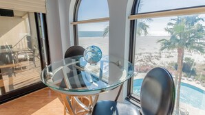 A room with a glass table and chairs, a view of the beach, and a pool.