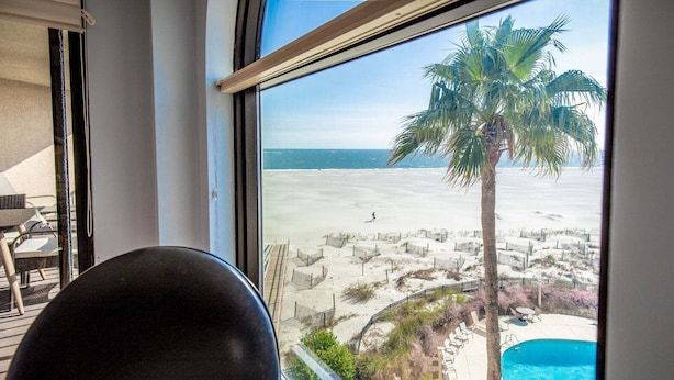 A room with a large window overlooking a beach, a pool, and a palm tree.