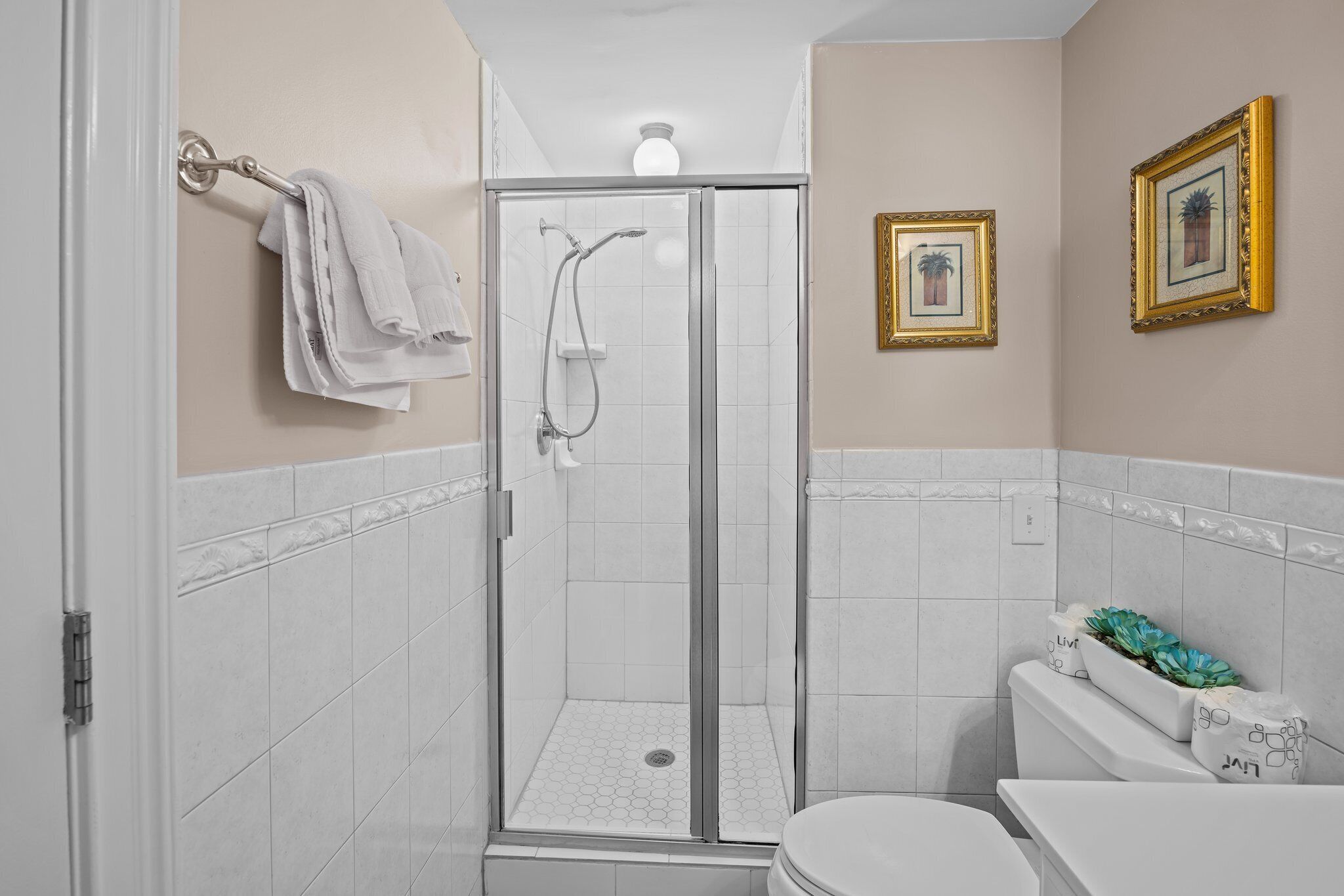 Un baño con ducha de vidrio, azulejos blancos, inodoro y cuadros en la pared.